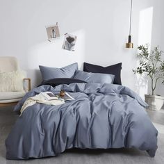 a bed with blue sheets and pillows in a white room next to a chair, potted plant and pictures on the wall