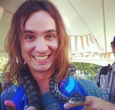 a man holding a snake in his right hand and smiling at the camera while another man looks on