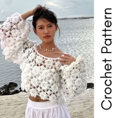 a woman standing on top of a beach next to the ocean wearing a white crochet sweater