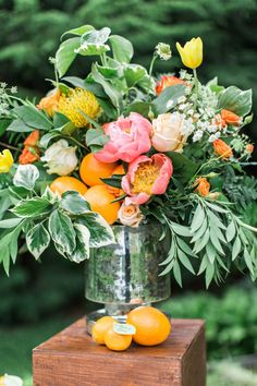 a vase filled with flowers and oranges on top of a wooden block next to trees