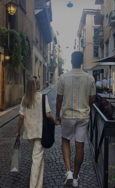 a man and woman holding hands walking down an alleyway with buildings in the background