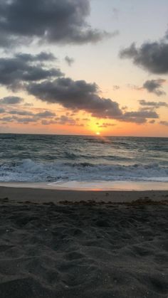 the sun is setting over the ocean with clouds in the sky and sand on the beach