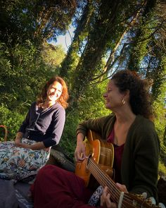 two women are sitting on the ground playing guitars and smiling at each other while another woman sits in the background