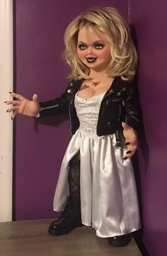 a doll dressed in white and black is standing on a purple wall with her hands out