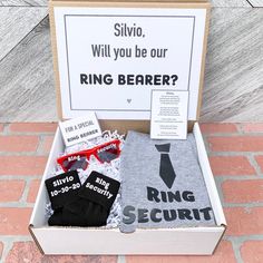 an open box with some items in it on a brick floor next to a sign that says, will you be our ring bearer?