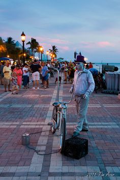 a man standing next to a bike on a sidewalk near the ocean at dusk with people walking around