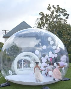 two girls in dresses are walking through a bubble ball on the grass with balloons floating around them