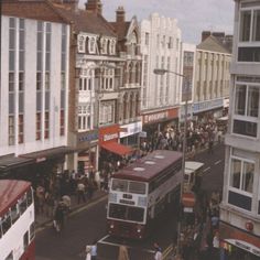 a busy city street with double decker buses and people walking on the sidewalk, in front of tall buildings