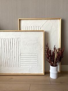 two white framed art pieces sitting on top of a wooden floor next to a vase filled with flowers