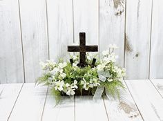 a cross with flowers in it sitting on a white wooden table next to a wall