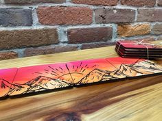 a piece of wood that has been painted with mountains and the sun on it, sitting on a wooden table