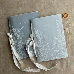 two blue book covers with white flowers on them and ribbon tied around the edges, sitting next to each other