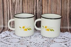 two white coffee mugs with yellow flowers on them sitting on a lace doily