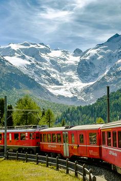 a red train traveling through a lush green hillside covered in snow capped mountain peaks and trees