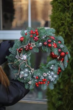 a woman holding a christmas wreath in front of a window with red and silver ornaments on it