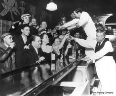 a group of men standing around a bar with drinks in front of them and one man reaching for a bowling ball