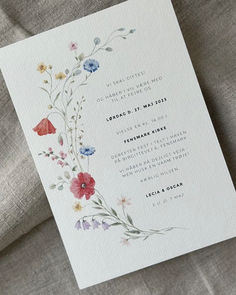 a wedding card with watercolor flowers on it sitting on top of a linen covered bed