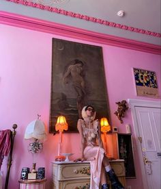 a woman sitting on top of a dresser in front of a pink wall with a painting above it