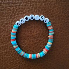 a beaded bracelet with the word cowgirl written in white and blue beads sits on a brown leather surface