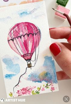 a hand holding a card with a hot air balloon on it and watercolors next to it