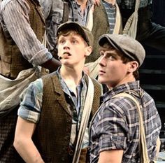 three men standing next to each other in front of a stage curtain and one man wearing a hat