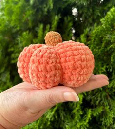 a hand holding a tiny crocheted pumpkin in front of some bushes and trees