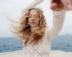 a woman with curly hair is holding her hands up in the air by the ocean