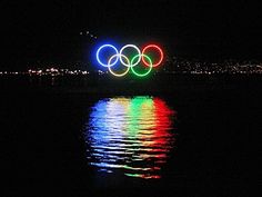 the olympic rings are lit up in the dark water at night with lights reflecting on the water