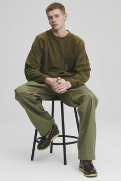 a young man sitting on top of a stool wearing green pants and a brown sweater