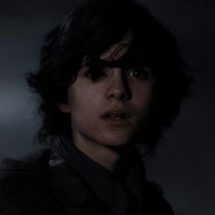 a man with black hair and dark clothes looks at the camera in a darkened room