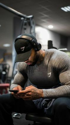 a man sitting in a chair with headphones on looking at his cell phone while wearing a hat