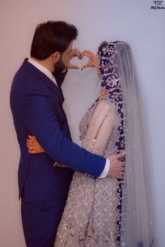 a man and woman standing next to each other in front of a white wall with a heart made out of their hands