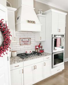 A wreath covered with red berries hangs above a warm wood floor. A Christmas sign hangs on the white subway tile backsplash above a white bowl on a black grill on the mottled brown counter on the white cabinets. Red kitchen accessories are strewn about the room...   Image: jk_farmhouselove