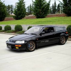 a black car parked in a parking lot next to some bushes and trees on the side of the road