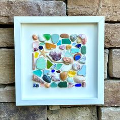 a white frame filled with lots of different colored sea glass and shells on top of a stone wall