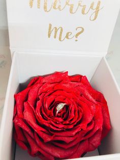 a red rose in a white box with the words marry me? written on it