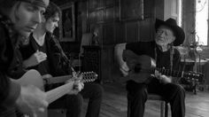 three people sitting in a room with guitars and one person holding an acoustic guitar while the other man plays