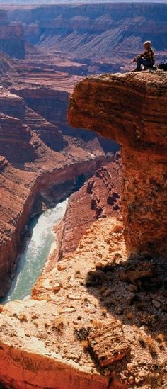 a man sitting on the edge of a cliff overlooking a river and canyon in the distance