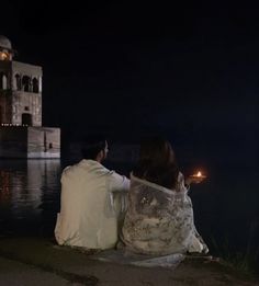 two people sitting on the edge of a body of water at night looking out to sea