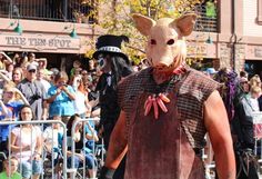 a man wearing a pig mask walking down the street in front of a large crowd