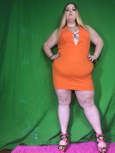 a woman in an orange dress poses for a photo on a pink rug with her hands on her hips