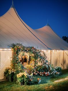 a large white tent with flowers and people standing in the doorway at night, surrounded by greenery