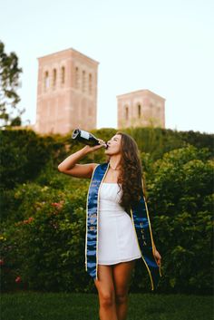 a woman in a white dress drinking from a bottle while wearing a blue and yellow graduation robe