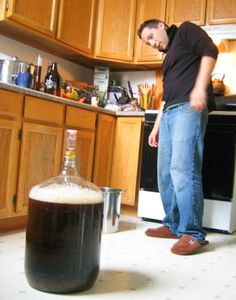 a man standing in a kitchen next to a large jug filled with beer and looking at the floor