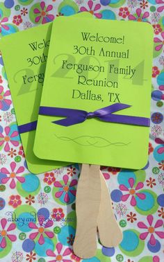 two green fan shaped fans sitting on top of a flowery tablecloth with purple ribbon