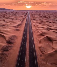 an empty road in the middle of desert with sun setting behind it and clouds overhead