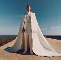 a man standing on top of a sandy beach next to the ocean wearing a white cape