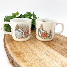 two coffee mugs sitting on top of a wooden board next to a plant and some leaves