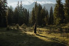 a woman standing on top of a grass covered field next to trees and mountains in the background