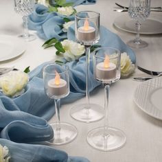 three candles are placed in wine glasses on a table with blue cloth and white flowers